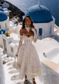 A woman wearing a strapless lace wedding gown with long sheer sleeves and a fitted corset-style bodice stands on white stone stairs in a Santorini village with blue-domed buildings and sea in the background. The dress is ivory/cream lace, and the scene includes visible text branding for Anna Elsie.