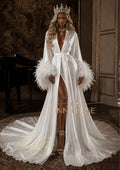 A woman models a luxurious white satin robe with dramatic feathered cuffs, a high thigh slit, and a matching train, styled with a silver crown in an elegant, dimly lit interior.